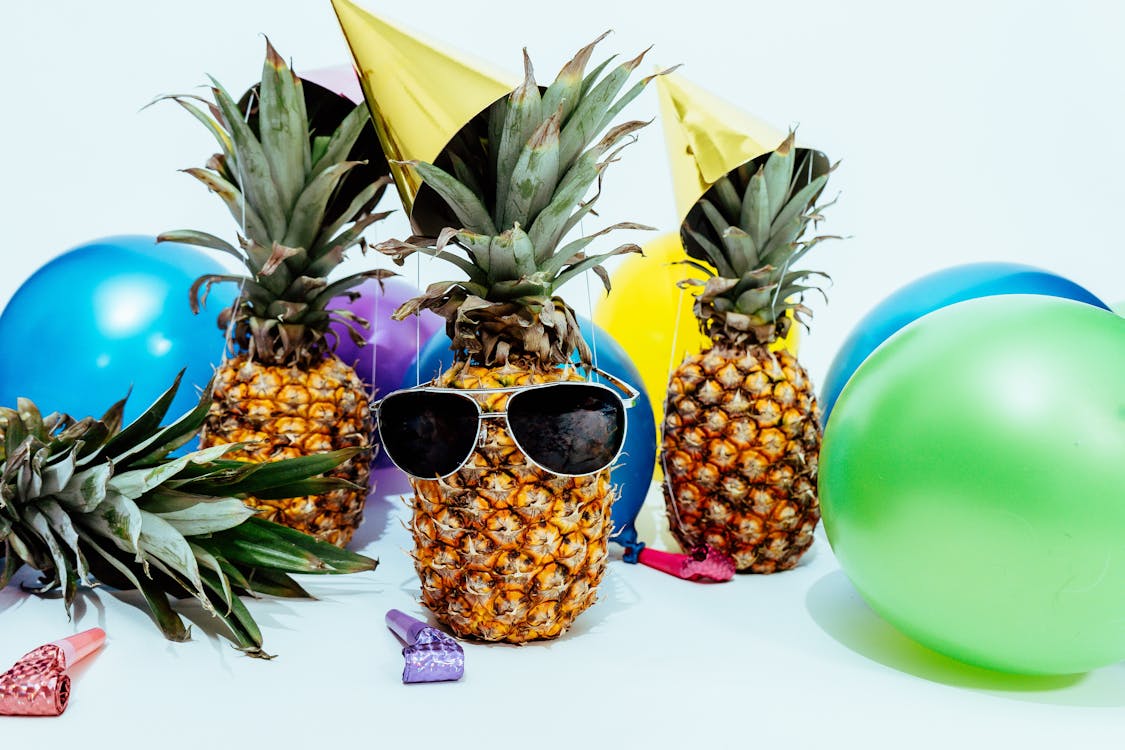 A pineapple wearing sunglasses wearing a gold party hat with multicolored balloons and party favors in the background