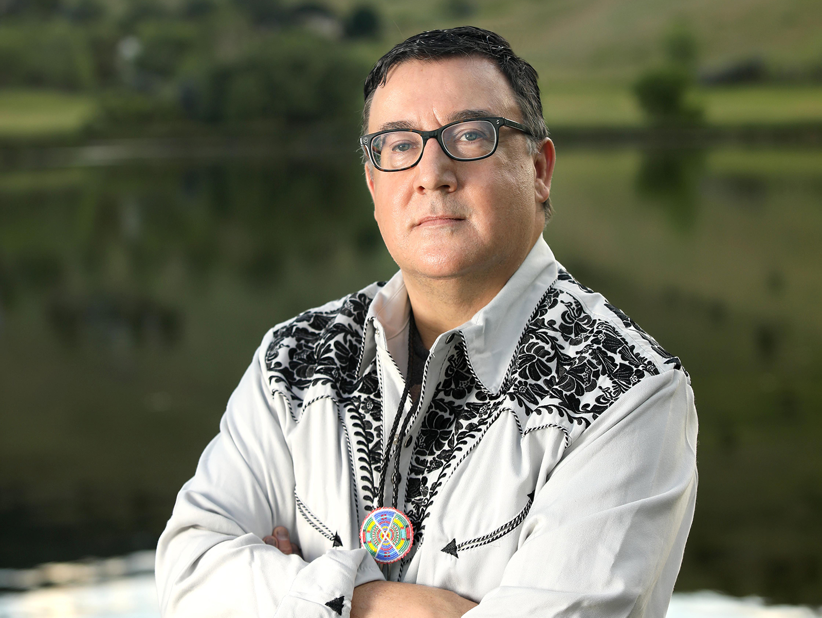 This image shows a man standing outdoors in front of a lake with a blurred background of greenery and hills. He is wearing glasses and a light-colored shirt with intricate black embroidery. His arms are crossed, and he has a serious expression on his face. He also wears a colorful medallion around his neck.