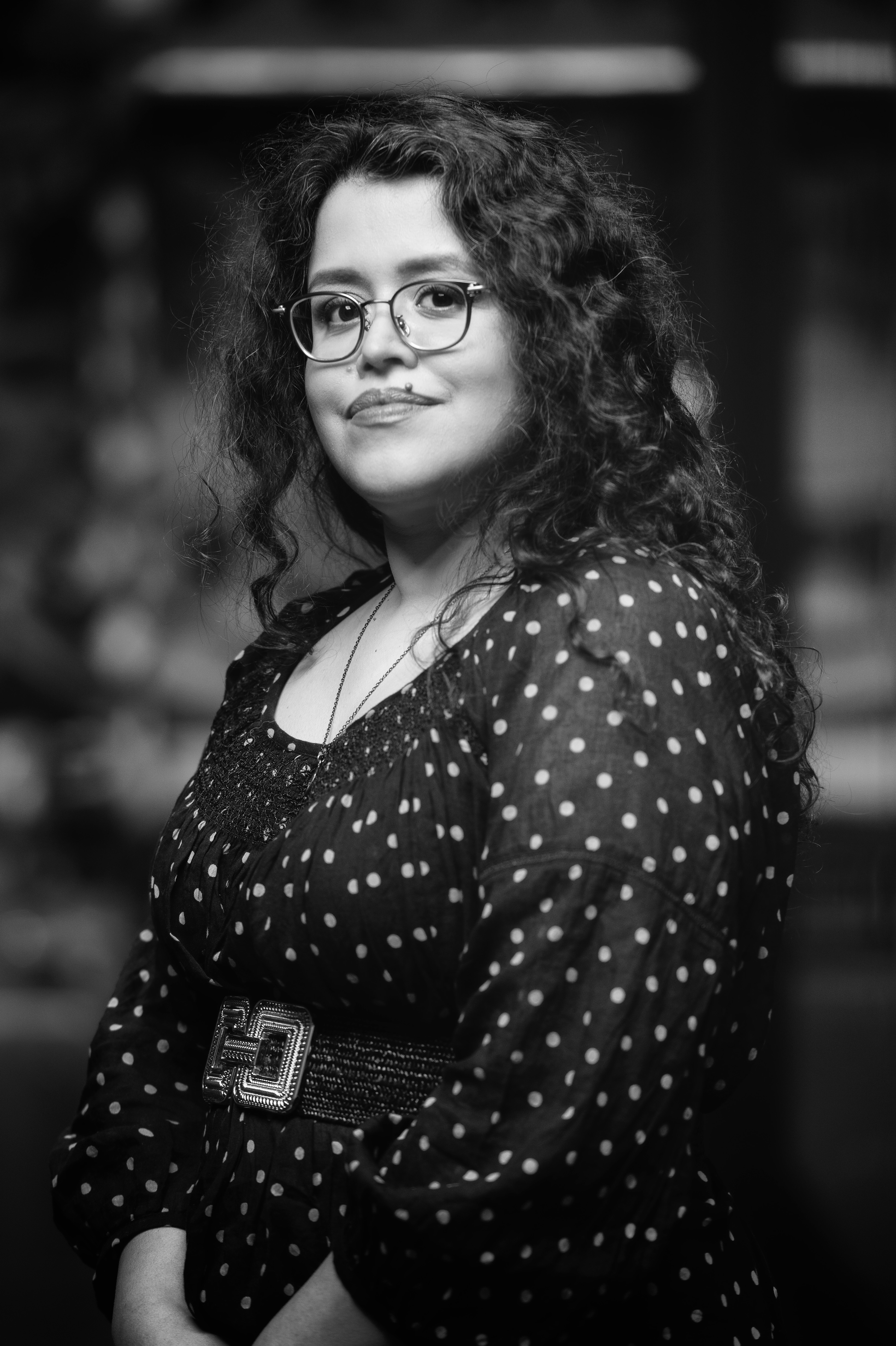  A grayscale portrait of a woman with curly hair, glasses, and a lip piercing. She is wearing a dark polka-dot dress with a wide, decorative belt. The background is softly blurred, drawing attention to her confident expression.