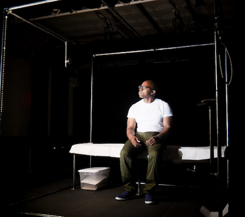 Photo of man on stage sitting on a bed which represents an incarcerated environment