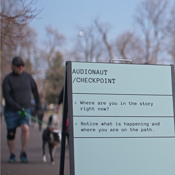 A sign reading “Audionaut / Checkpoint” stands along a park path with reflection prompts for participants, while a person walks a dog in the background.