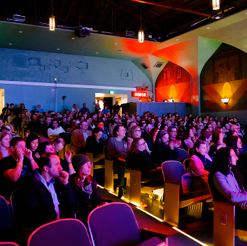 Audience members fill a large auditorium during a live library event, seated and facing the stage under colorful theatrical lighting.