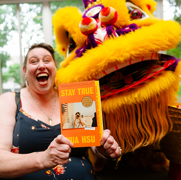 A smiling person holds up the book Stay True by Hua Hsu while posing beside a bright yellow and pink lion dance costume inside Parkview event space in Central Library.