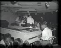 A black and white photo of the Rainbow Ballroom stage. There is a Black man playing piano and a band guddled together holding string instruments.